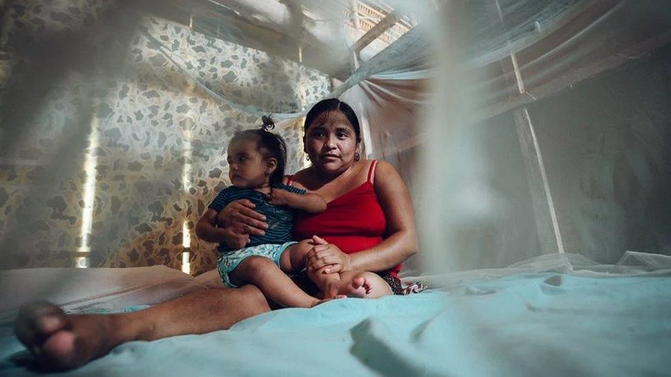 An adult and child sitting together on a bed inside mosquito netting, highlighting family protection from maleria.