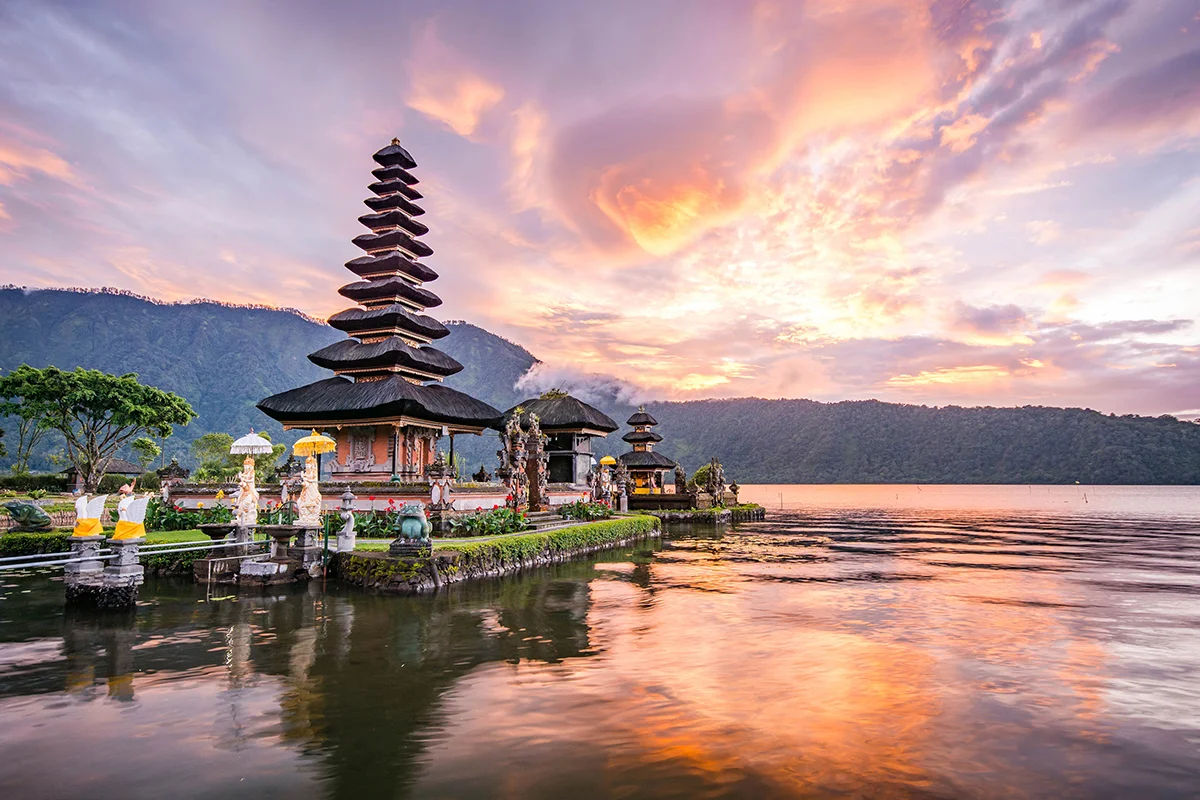 Lakeside temple with tiered pagoda roof at sunset, reflected in calm water with mountains in the background.