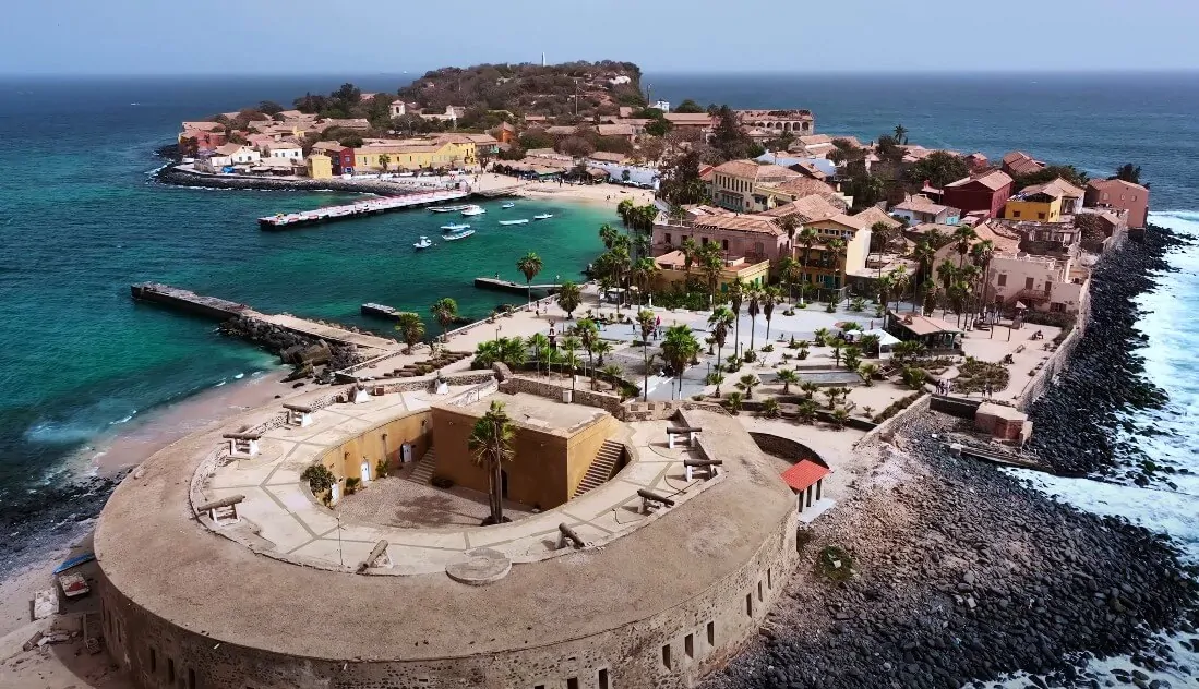 Aerial view of a coastal island town with colorful buildings, a harbor, and a historic fort by the sea.