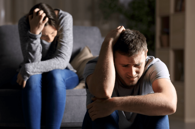 Two people indoors sitting with heads in hands, conveying a somber reflective mood.