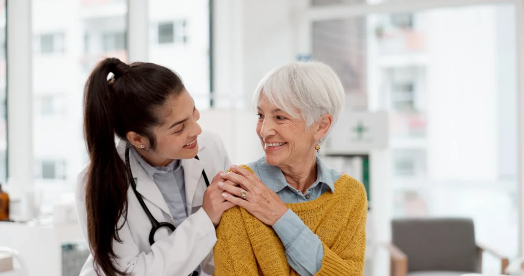 Doctor placing hands on older patient’s shoulders in a bright medical setting, symbolising care and support.
