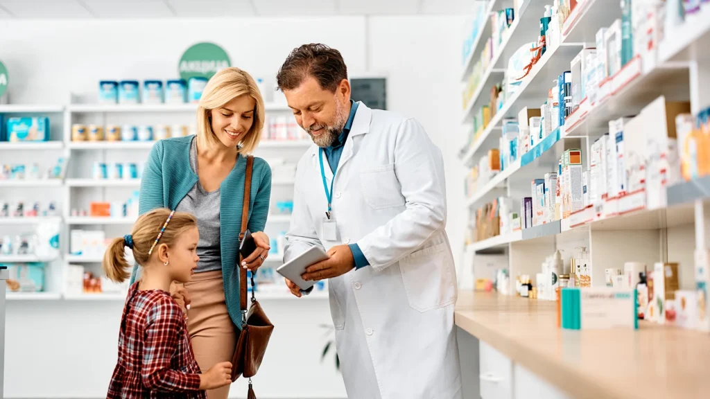 Pharmacist showing information on a tablet to an adult and child inside a bright pharmacy.