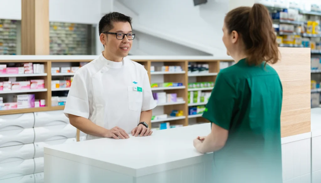 Pharmacist in uniform assisting customer across counter in a clean, organised pharmacy.