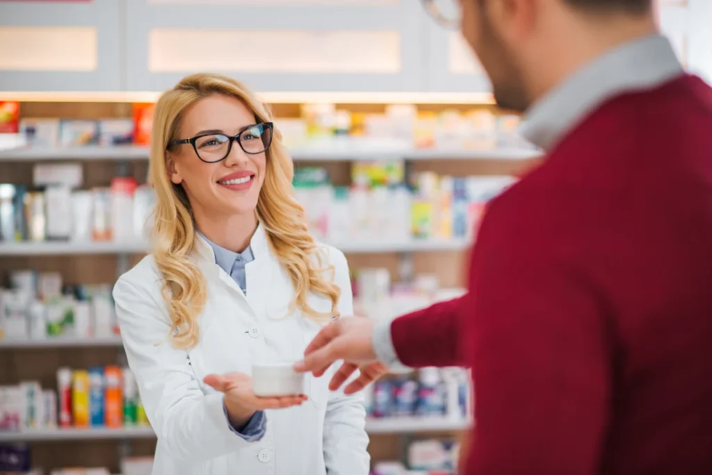 A pharmacist in a white lab coat hands a medication container to a customer at Yatton Pharmacy, Yatton, Bristol.
