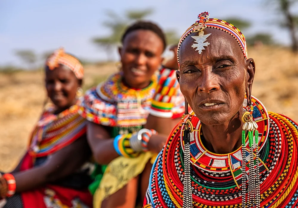A person in vibrant traditional beadwork and jewelry stands in focus, with two similarly adorned individuals behind them in a semi-arid outdoor setting.