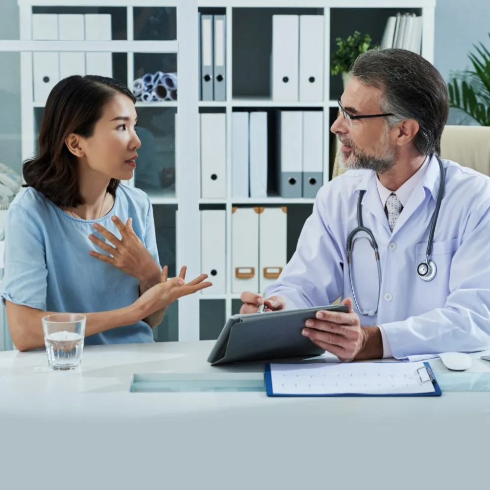 A healthcare professional in a white coat with a stethoscope consults a patient across a desk, holding a clipboard, at Yatton Pharmacy, Yatton, Bristol.