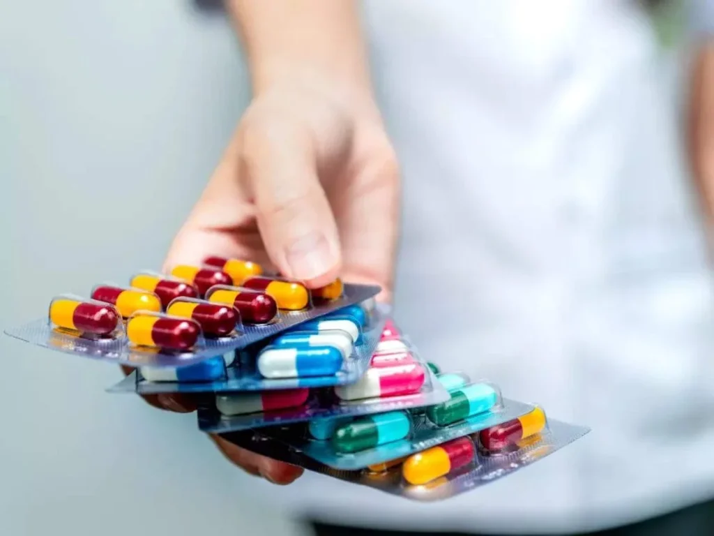 A tipped brown medicine bottle spills assorted tablets beside blister packs of capsules and a syringe on a wooden surface.
