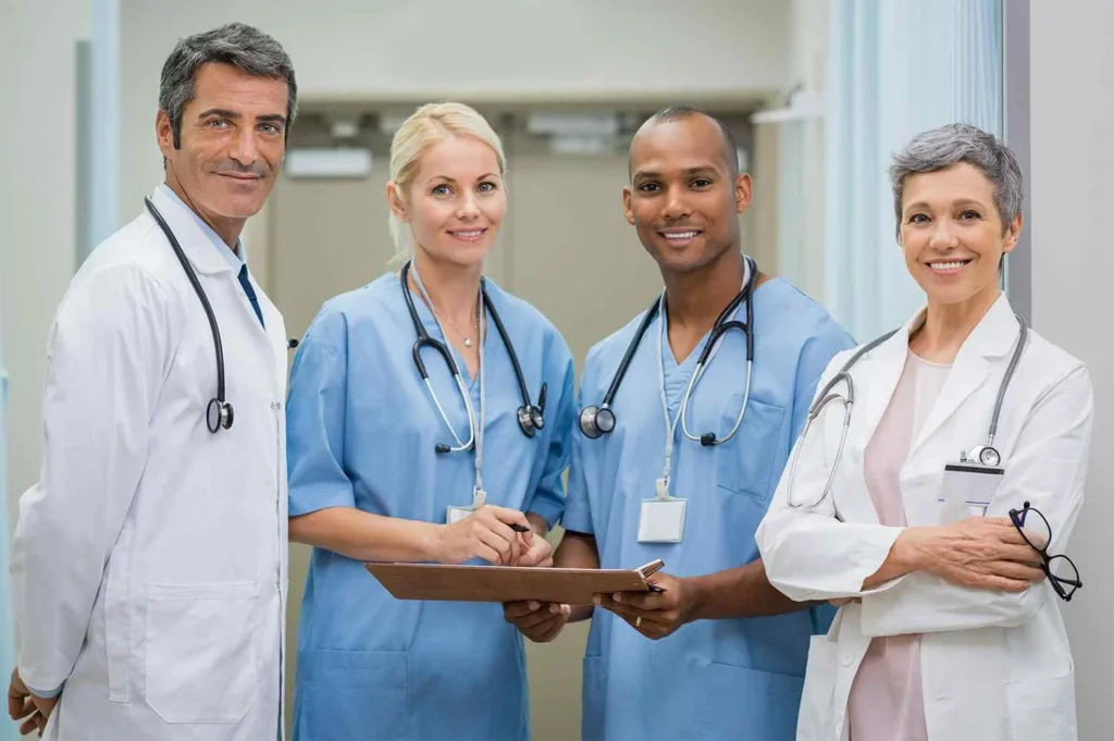 Four healthcare professionals two in white lab coats with stethoscopes and two in light blue scrubs stand together in a clinical hallway, collaborating on patient care at Yatton Pharmacy, Yatton, Bristol.