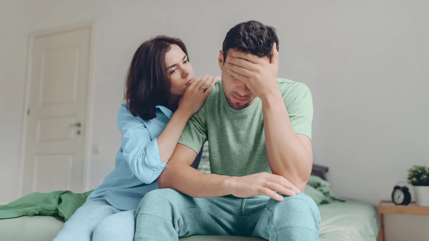 Two people sitting on a bed, one whispering while the other looks down with hand partly covering face.