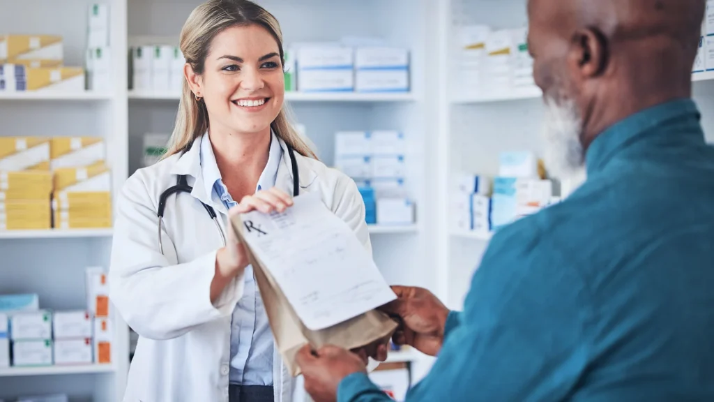 A smiling female pharmacist of Yatton Pharmacy handing a prescription bag and document to an older male customer in Yatton, Bristol.
