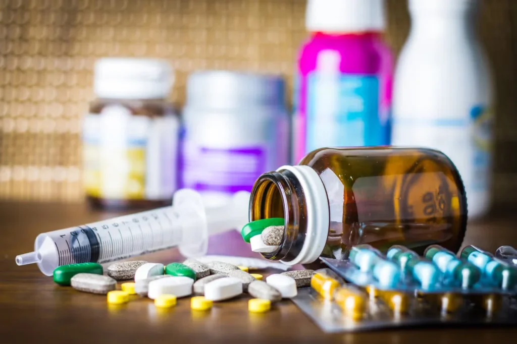 A tipped brown medicine bottle spills assorted tablets beside blister packs of capsules and a syringe on a wooden surface.