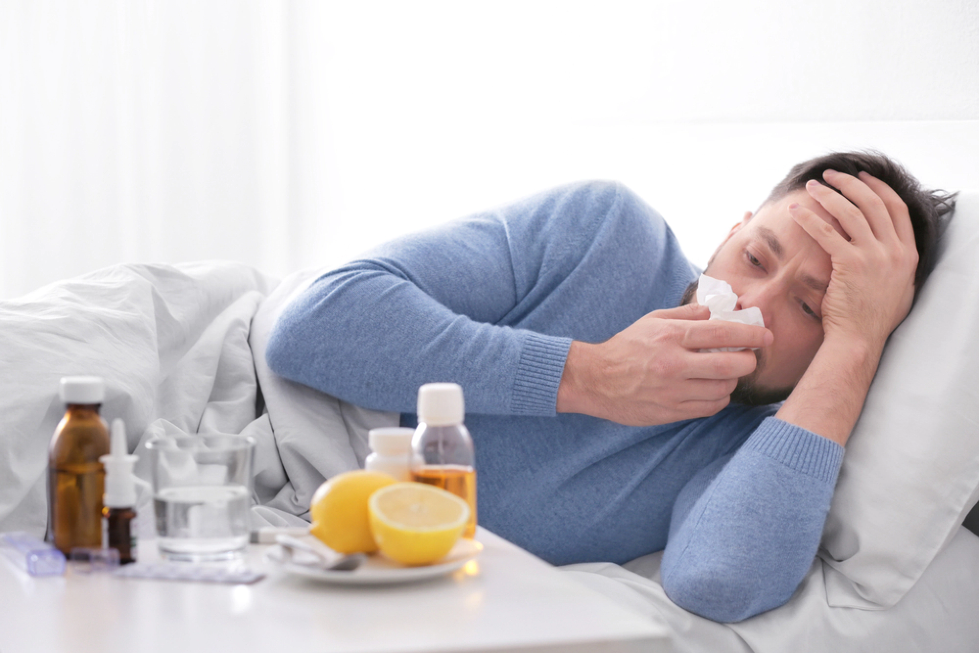 A person lies in bed appearing unwell, holding a tissue and surrounded by medicine bottles, tablets, and a sliced lemon Yatton, Bristol.