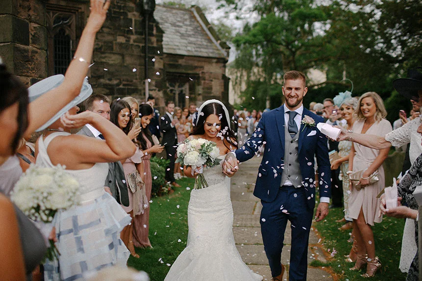 A newlywed couple walks joyfully down a path outside a stone church as guests throw confetti Yatton, Bristol.